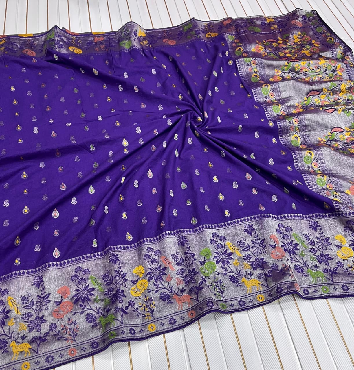 A purple Banarasi silk saree or fabric piece with small metallic motifs and a heavily embroidered floral border, laid flat on a white slatted surface.
